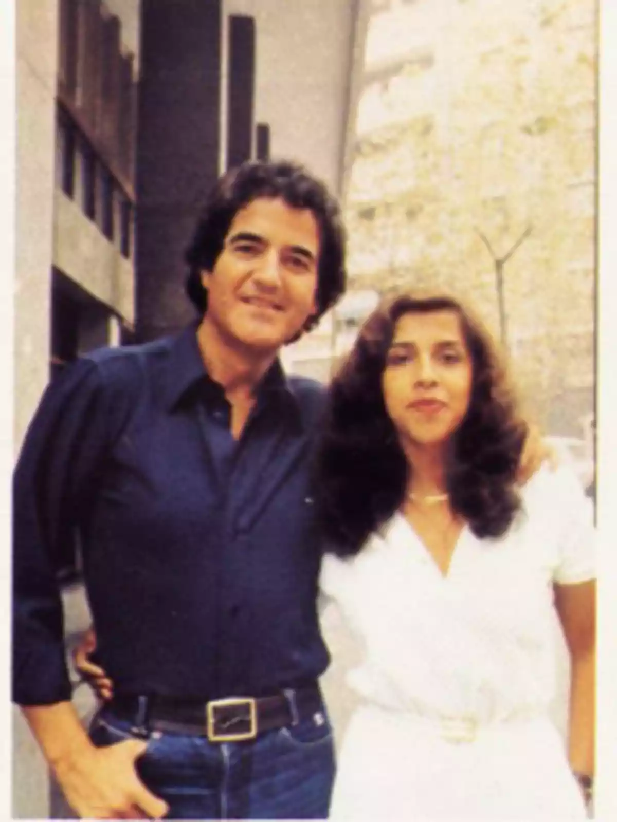 Una pareja posa abrazada en la calle, el hombre viste camisa azul y jeans, la mujer lleva un vestido blanco y ambos sonríen frente a un edificio. Una pareja posa abrazada en la calle, el hombre viste camisa azul y jeans, la mujer lleva un vestido blanco y ambos sonríen frente a un edificio.