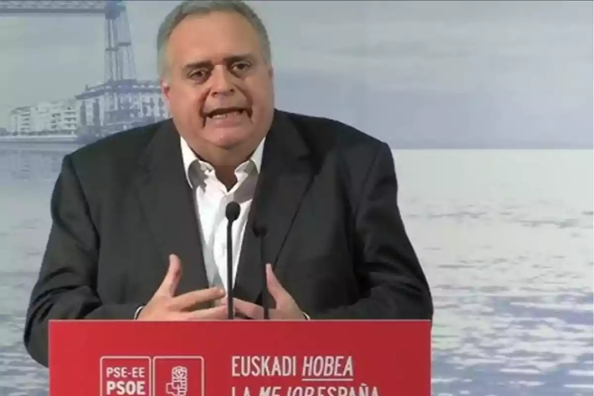 Hombre de cabello canoso y traje oscuro hablando en un atril rojo con logotipos del PSOE y el texto Euskadi HOBEA LA MEJOR ESPAÑA, con un fondo de paisaje urbano y agua. Hombre de cabello canoso y traje oscuro hablando en un atril rojo con logotipos del PSOE y el texto Euskadi HOBEA LA MEJOR ESPAÑA, con un fondo de paisaje urbano y agua.