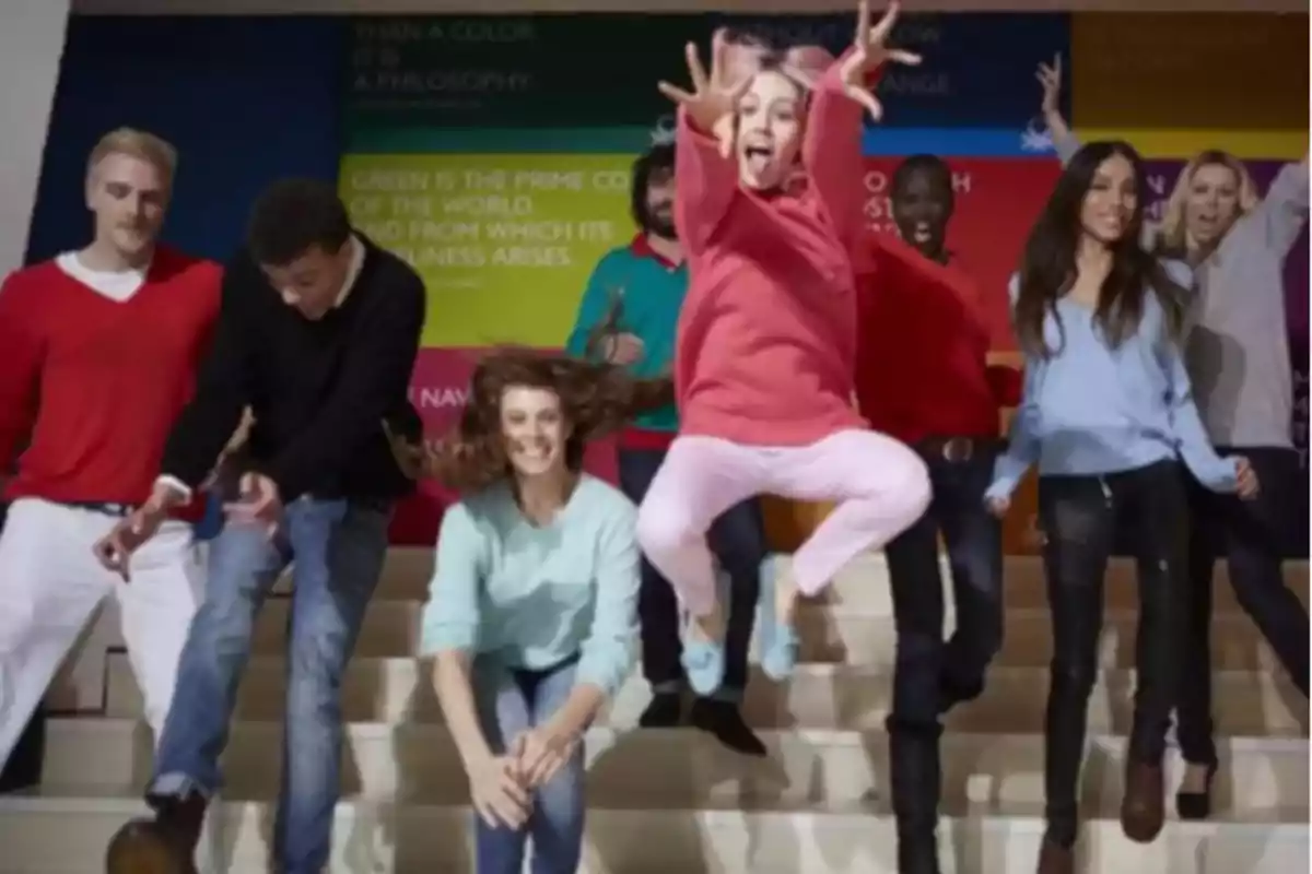 Un grupo de personas jóvenes sonríe y salta en unas escaleras mientras una niña en primer plano salta con entusiasmo y saca la lengua Un grupo de personas jóvenes sonríe y salta en unas escaleras mientras una niña en primer plano salta con entusiasmo y saca la lengua