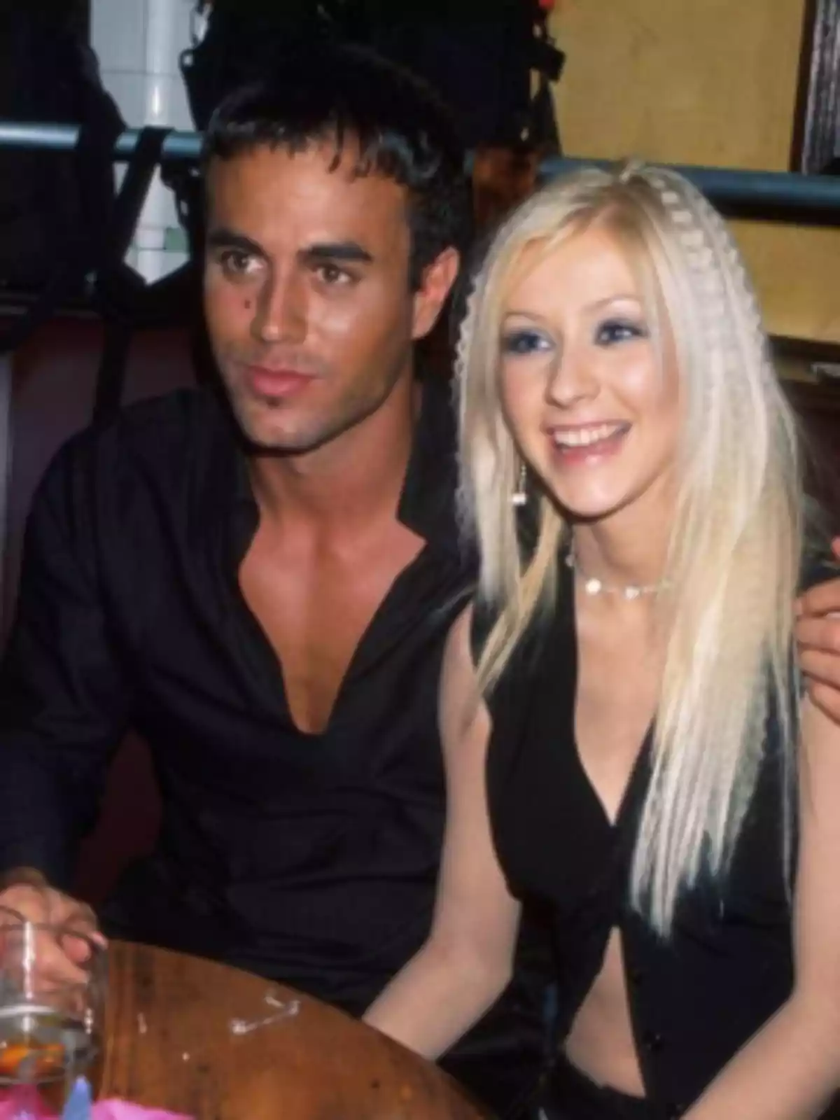 Dos personas jóvenes sentadas juntas en una mesa, ambas visten de negro y sonríen mientras miran hacia el frente Dos personas jóvenes sentadas juntas en una mesa, ambas visten de negro y sonríen mientras miran hacia el frente