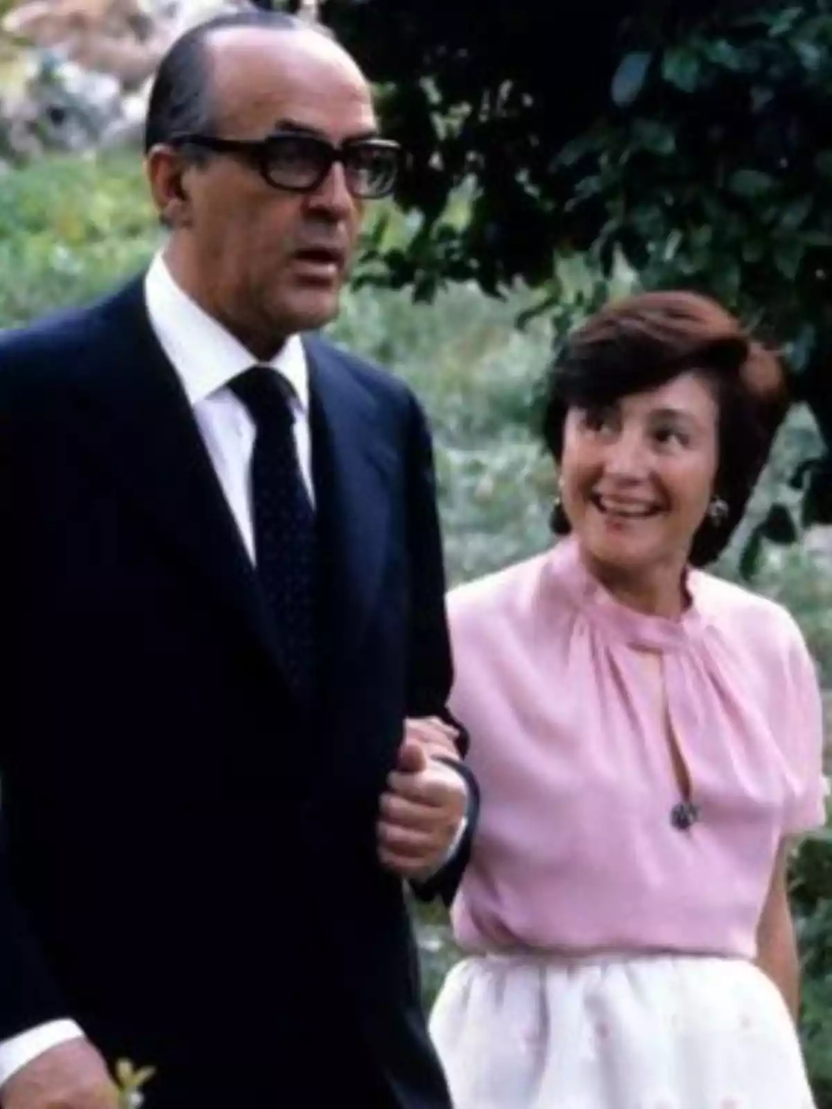 Pareja caminando al aire libre, el hombre lleva traje oscuro y gafas, la mujer viste blusa rosa claro y falda blanca, ambos parecen conversar y ella sonríe Pareja caminando al aire libre, el hombre lleva traje oscuro y gafas, la mujer viste blusa rosa claro y falda blanca, ambos parecen conversar y ella sonríe
