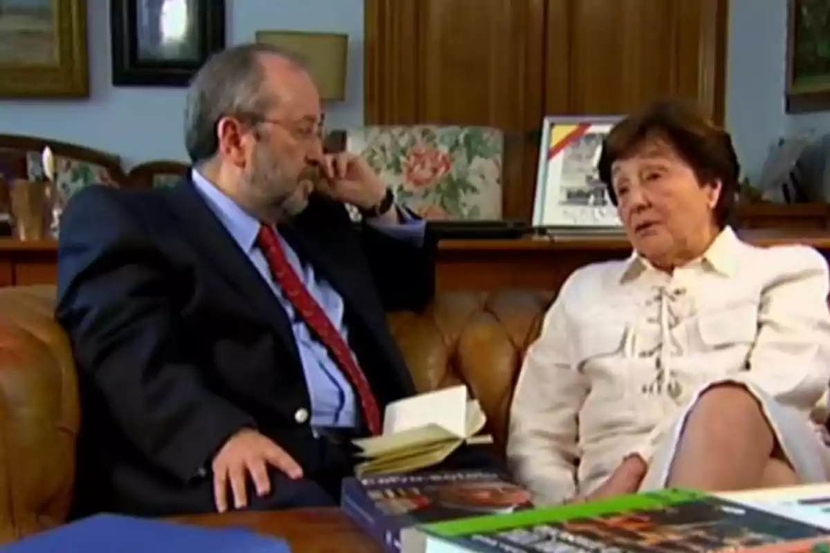 Un hombre con traje y una mujer mayor conversan sentados en un sofá de cuero en una sala con libros y cuadros al fondo Un hombre con traje y una mujer mayor conversan sentados en un sofá de cuero en una sala con libros y cuadros al fondo
