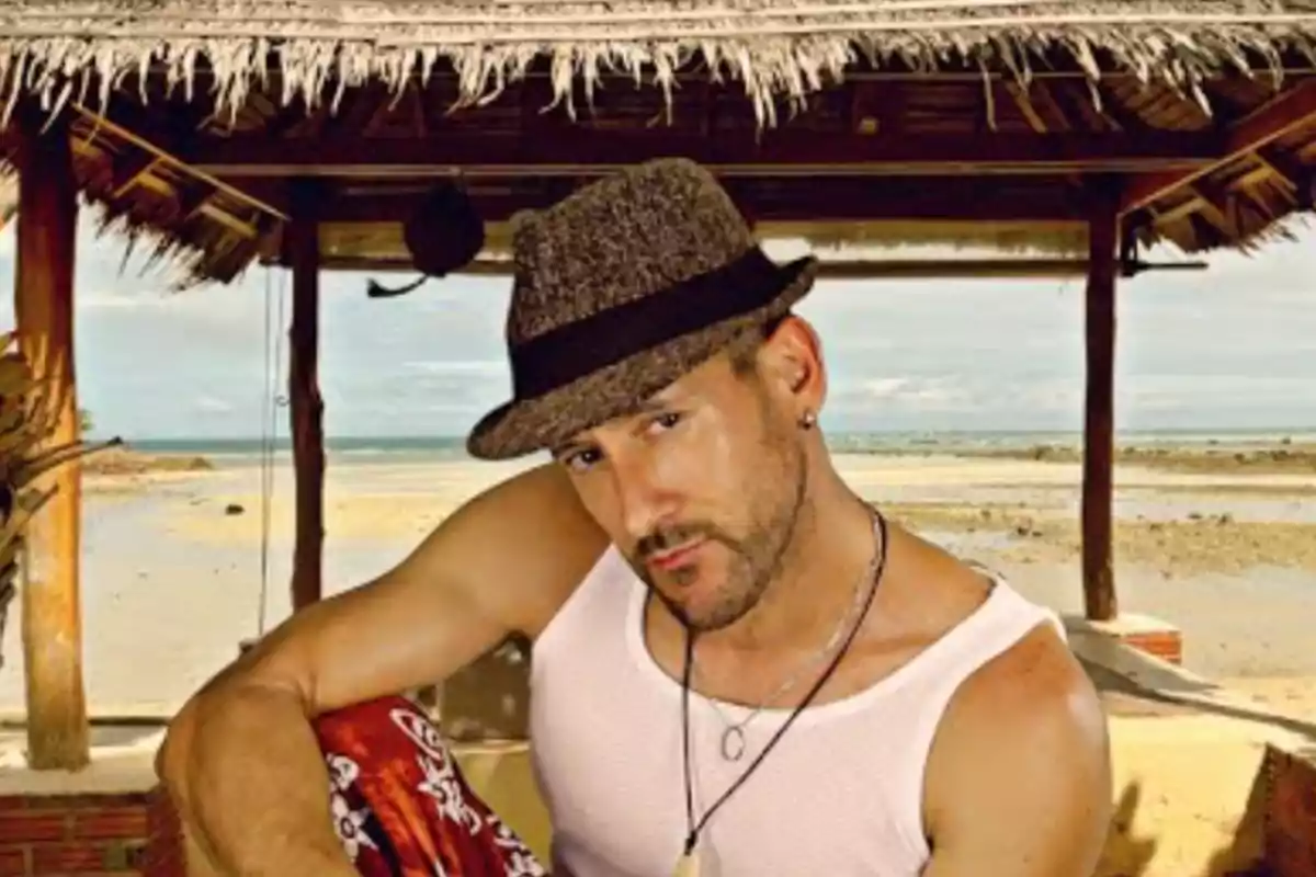 Hombre con sombrero y camiseta sin mangas posando bajo una palapa en la playa