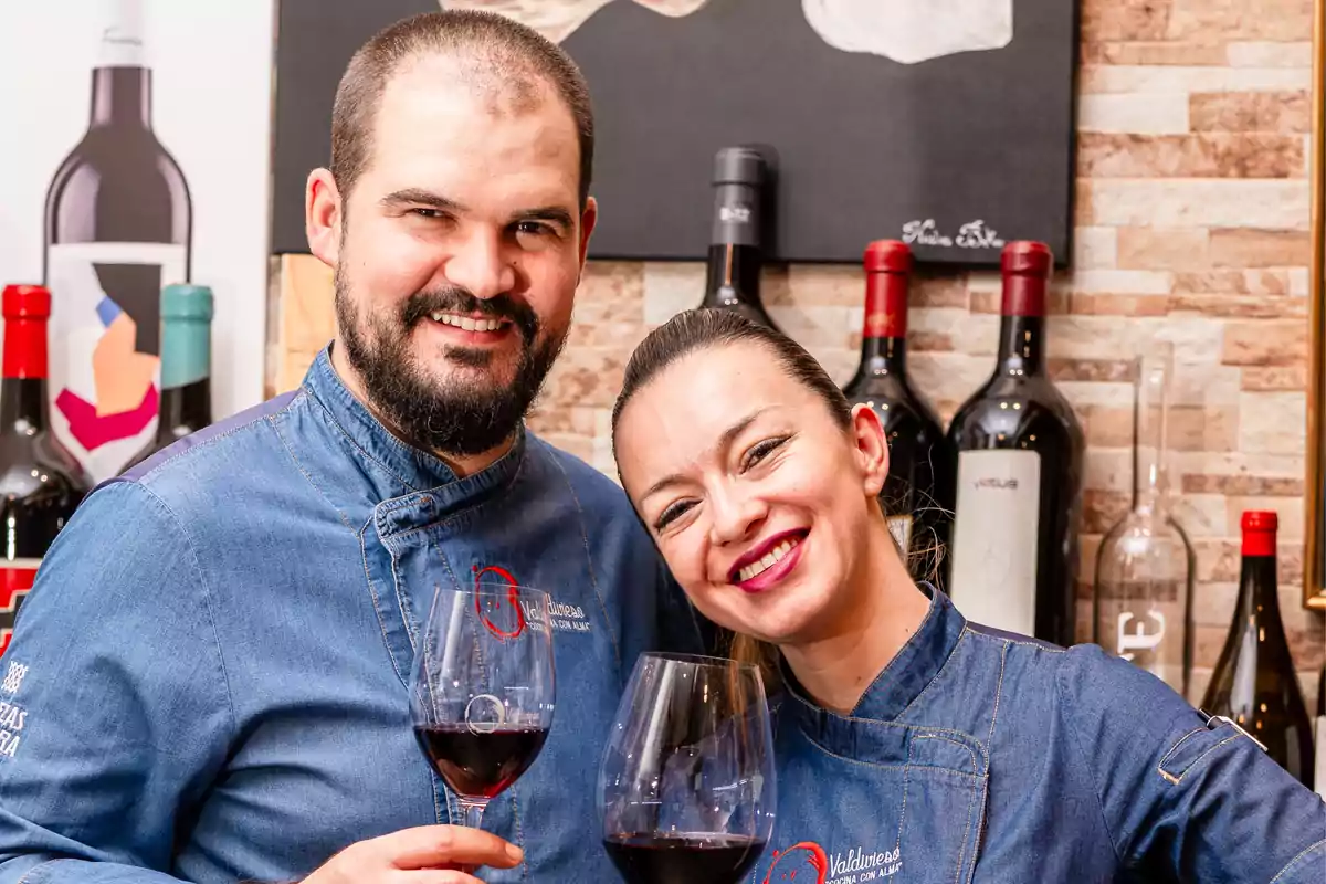 Dos personas sonrientes con copas de vino tinto, vistiendo chaquetas de chef azules, posan frente a una pared decorada con botellas de vino. Dos personas sonrientes con copas de vino tinto, vistiendo chaquetas de chef azules, posan frente a una pared decorada con botellas de vino.