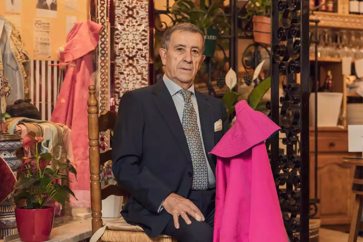 Hombre mayor con traje oscuro y corbata sentado en una silla de madera en un ambiente decorado con plantas y objetos tradicionales sosteniendo una capa de color rosa brillante Hombre mayor con traje oscuro y corbata sentado en una silla de madera en un ambiente decorado con plantas y objetos tradicionales sosteniendo una capa de color rosa brillante