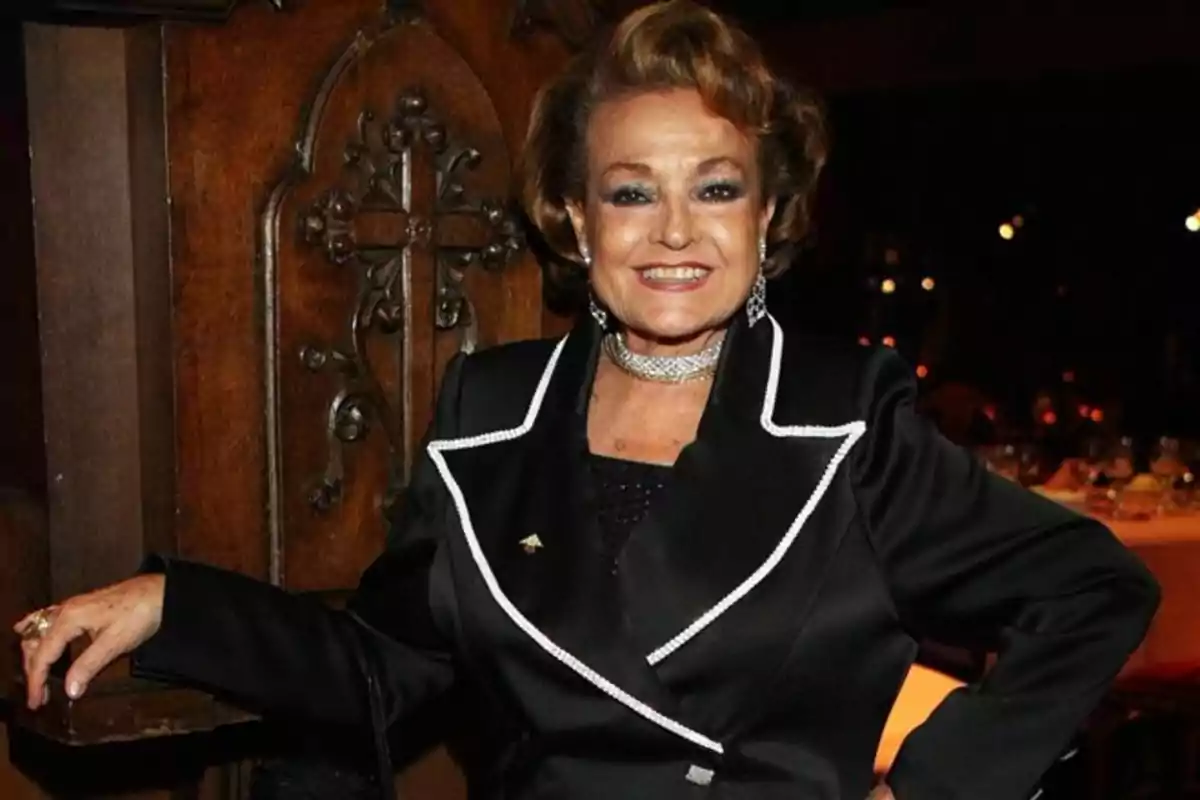 Mujer mayor sonriente con saco negro elegante y joyas, posando en un lugar con decoración de madera y luz tenue Mujer mayor sonriente con saco negro elegante y joyas, posando en un lugar con decoración de madera y luz tenue