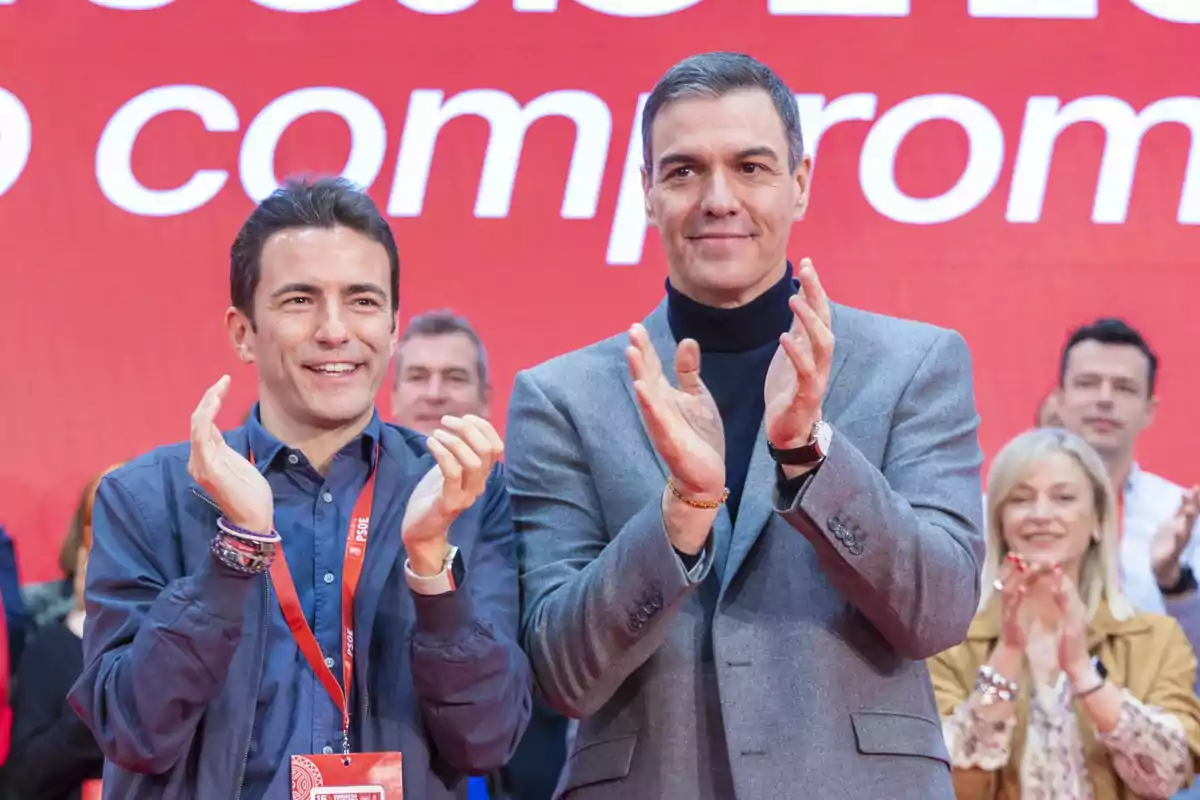Dos hombres aplauden sonrientes en un evento con fondo rojo y personas al fondo también aplaudiendo