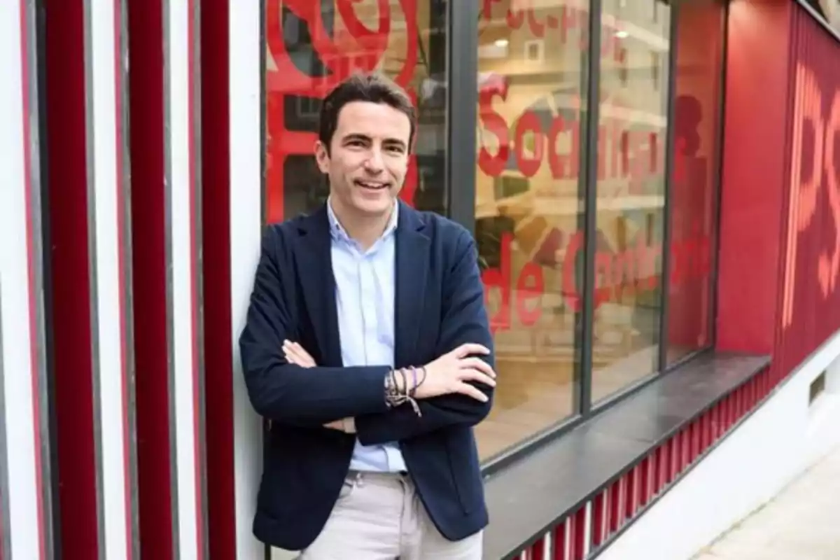 Hombre de cabello corto y oscuro con chaqueta azul y camisa clara posando sonriente con los brazos cruzados frente a un edificio con paredes rojas y ventanas grandes Hombre de cabello corto y oscuro con chaqueta azul y camisa clara posando sonriente con los brazos cruzados frente a un edificio con paredes rojas y ventanas grandes