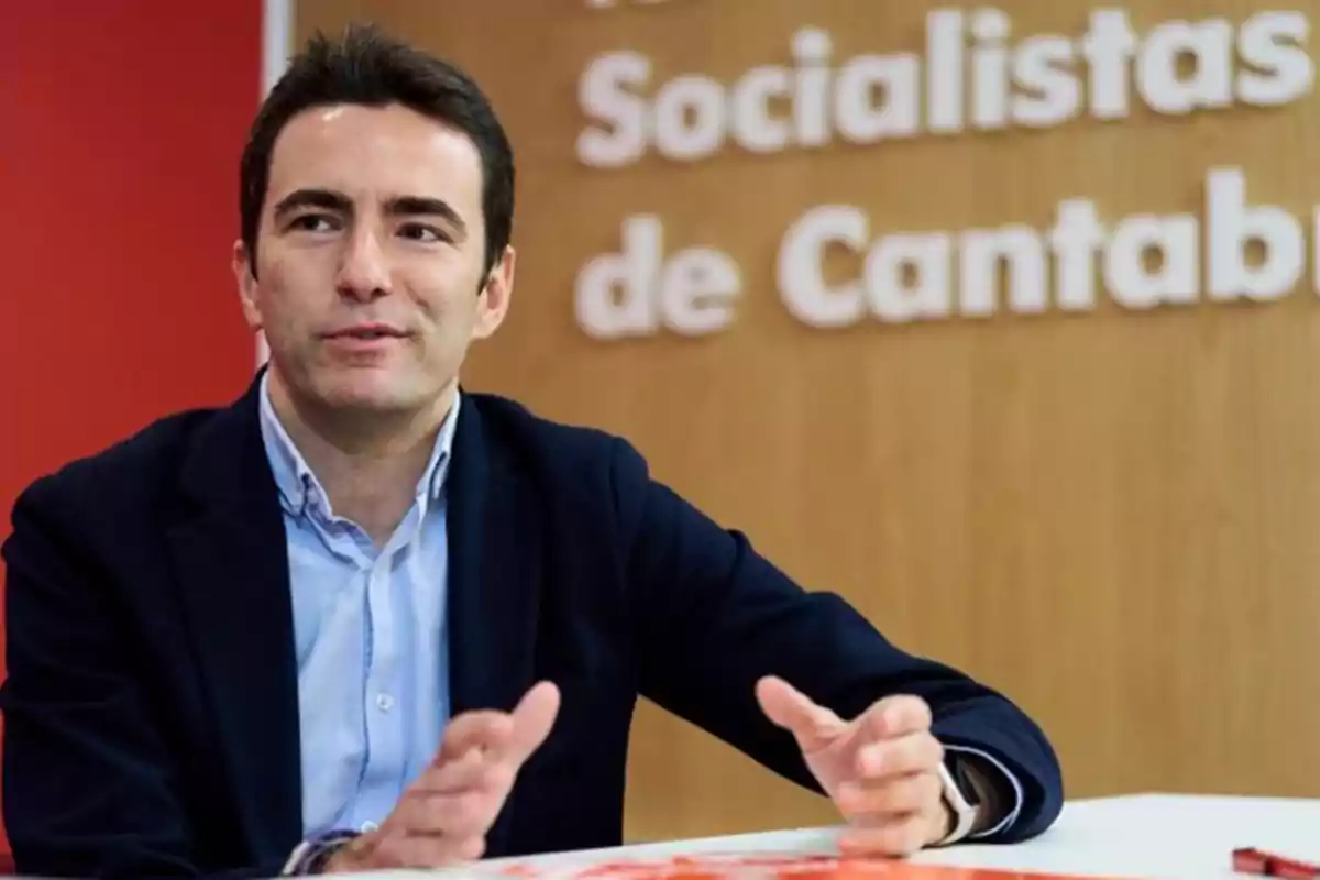 Hombre joven de cabello oscuro con chaqueta azul y camisa clara hablando y gesticulando con las manos frente a un fondo con letras blancas que dicen Socialistas de Cantabria Hombre joven de cabello oscuro con chaqueta azul y camisa clara hablando y gesticulando con las manos frente a un fondo con letras blancas que dicen Socialistas de Cantabria