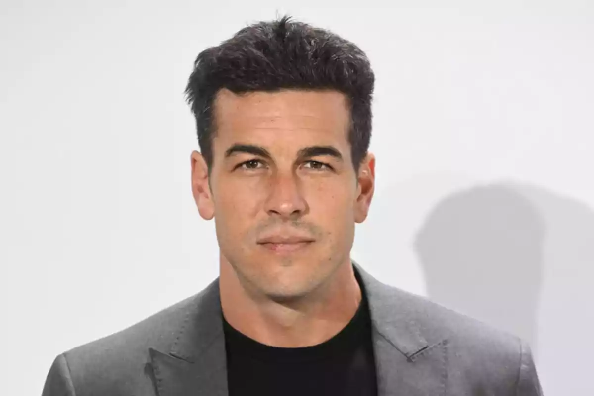 Hombre de cabello oscuro con saco gris y camiseta negra posando frente a un fondo blanco