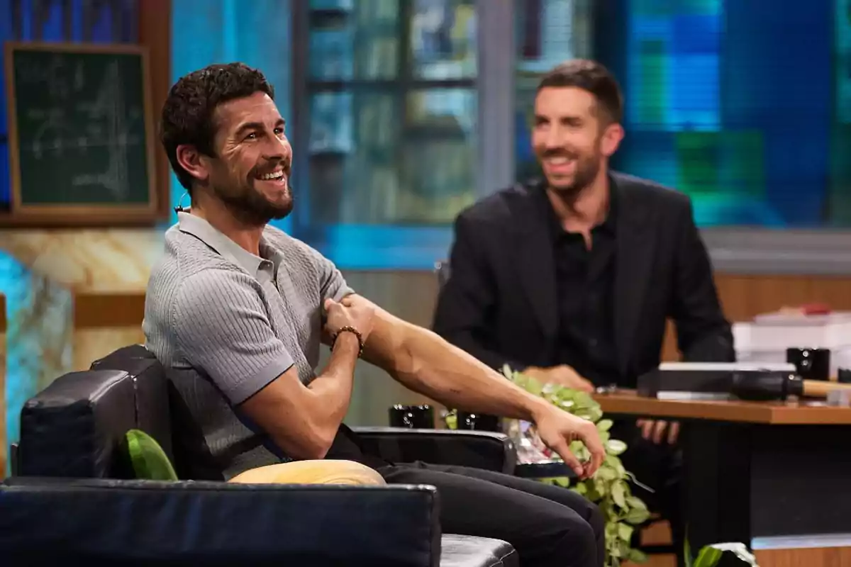 Dos hombres sonríen y conversan en un set de televisión con ambiente relajado y decorado con plantas y una pizarra al fondo Dos hombres sonríen y conversan en un set de televisión con ambiente relajado y decorado con plantas y una pizarra al fondo