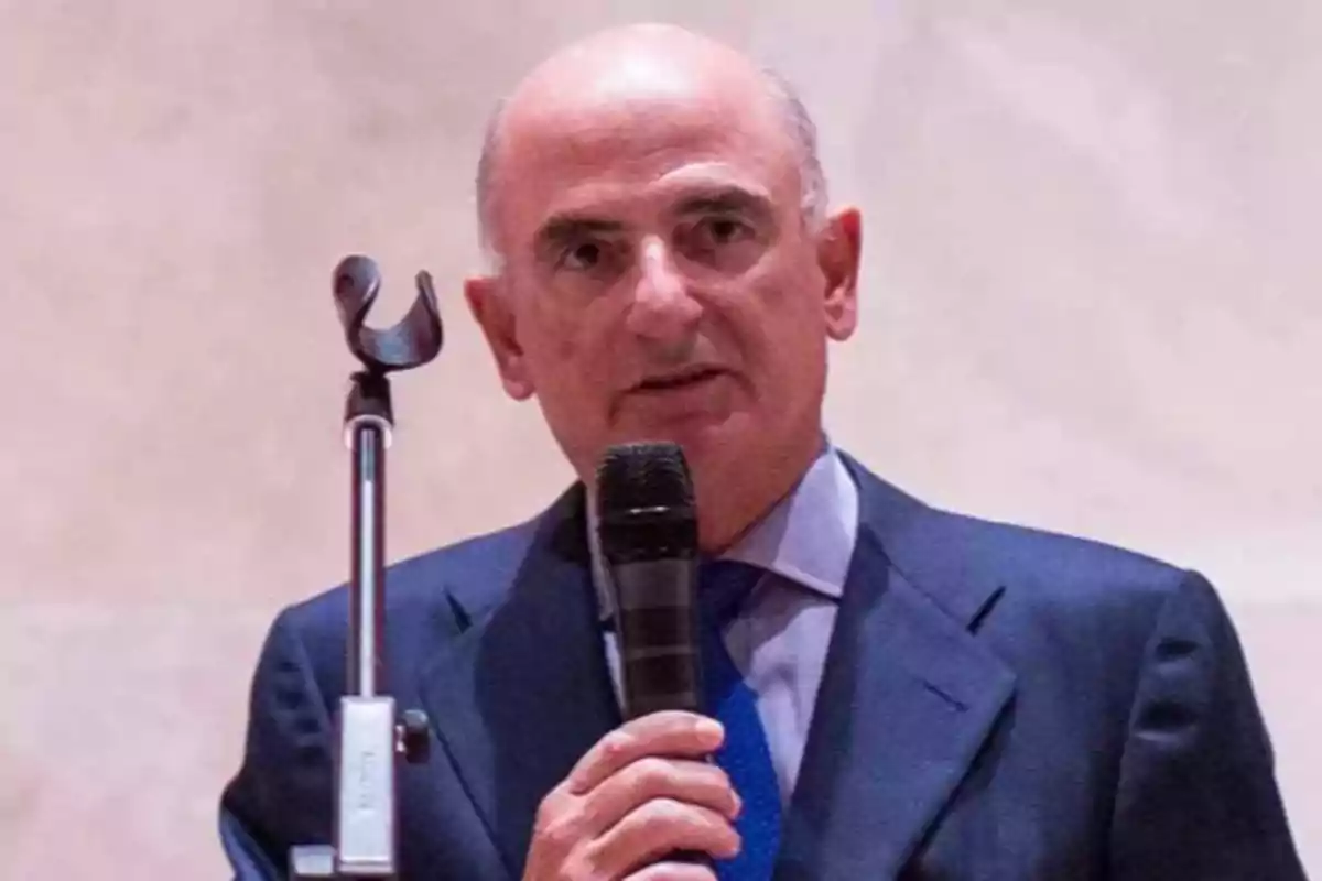 Hombre calvo con traje azul y corbata azul hablando por micrófono frente a un fondo claro