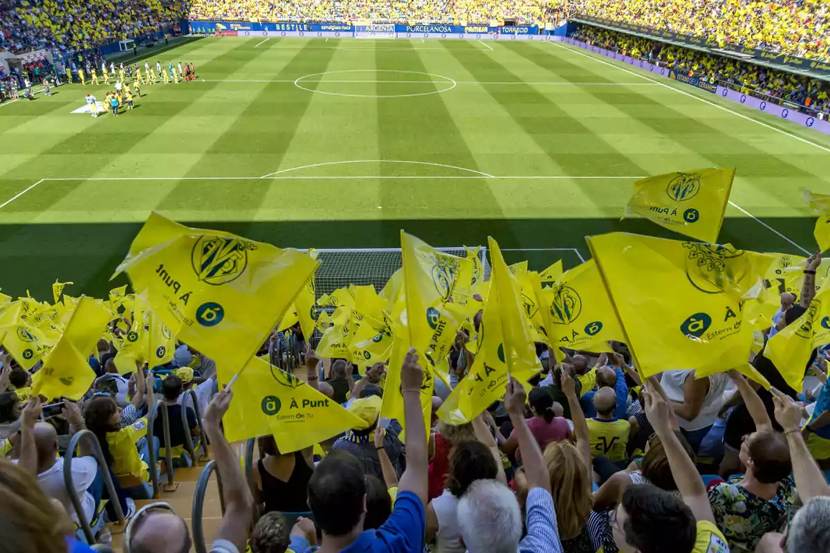 Aficionados en un estadio de fútbol ondeando banderas amarillas mientras los equipos salen al campo Aficionados en un estadio de fútbol ondeando banderas amarillas mientras los equipos salen al campo