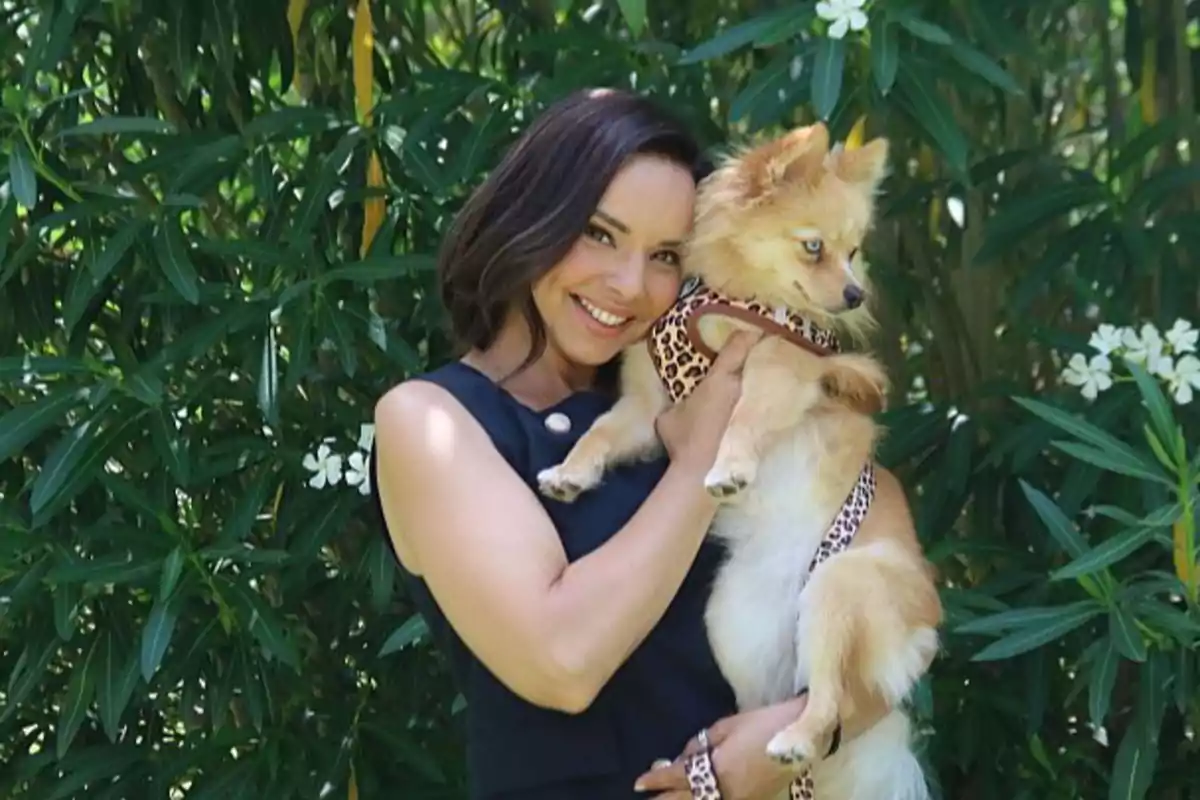 Mujer sonriente con cabello oscuro sostiene a un perro pequeño de pelaje claro y ojos azules, ambos posando frente a un arbusto verde con flores blancas Mujer sonriente con cabello oscuro sostiene a un perro pequeño de pelaje claro y ojos azules, ambos posando frente a un arbusto verde con flores blancas