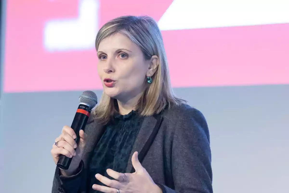 Mujer rubia hablando en público mientras sostiene un micrófono y gesticula con la mano frente a un fondo borroso de colores claros Mujer rubia hablando en público mientras sostiene un micrófono y gesticula con la mano frente a un fondo borroso de colores claros