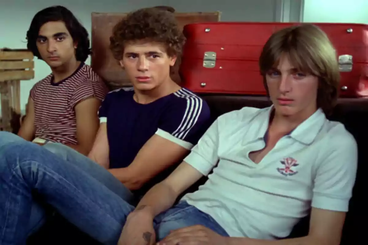 Tres jóvenes sentados en un sofá con expresión seria y maletas rojas al fondo Tres jóvenes sentados en un sofá con expresión seria y maletas rojas al fondo
