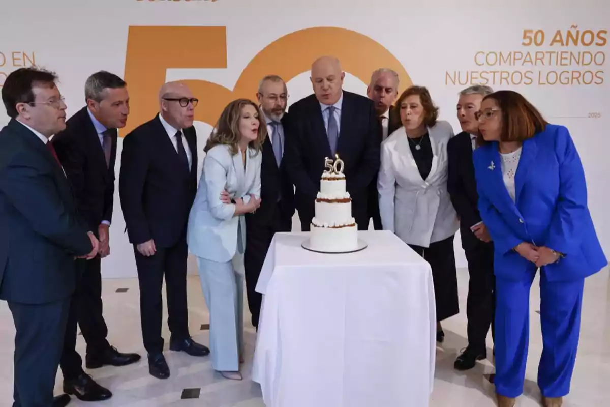 Un grupo de personas elegantemente vestidas sopla las velas de un pastel de tres pisos con el número 50 en la parte superior, celebrando un aniversario en un ambiente corporativo. Un grupo de personas elegantemente vestidas sopla las velas de un pastel de tres pisos con el número 50 en la parte superior, celebrando un aniversario en un ambiente corporativo.