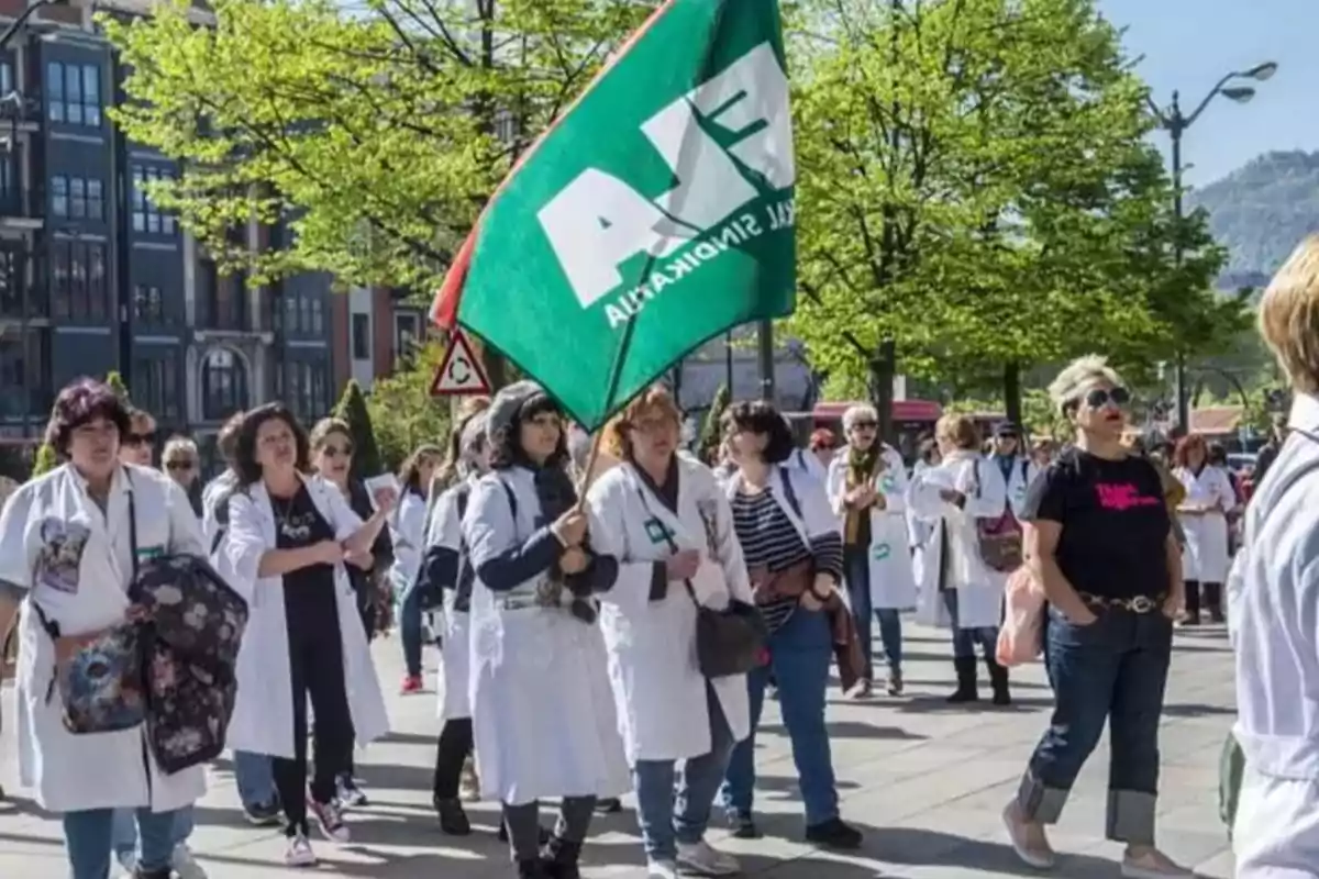 Grupo de personas con batas blancas marchando al aire libre mientras una de ellas sostiene una bandera verde Grupo de personas con batas blancas marchando al aire libre mientras una de ellas sostiene una bandera verde