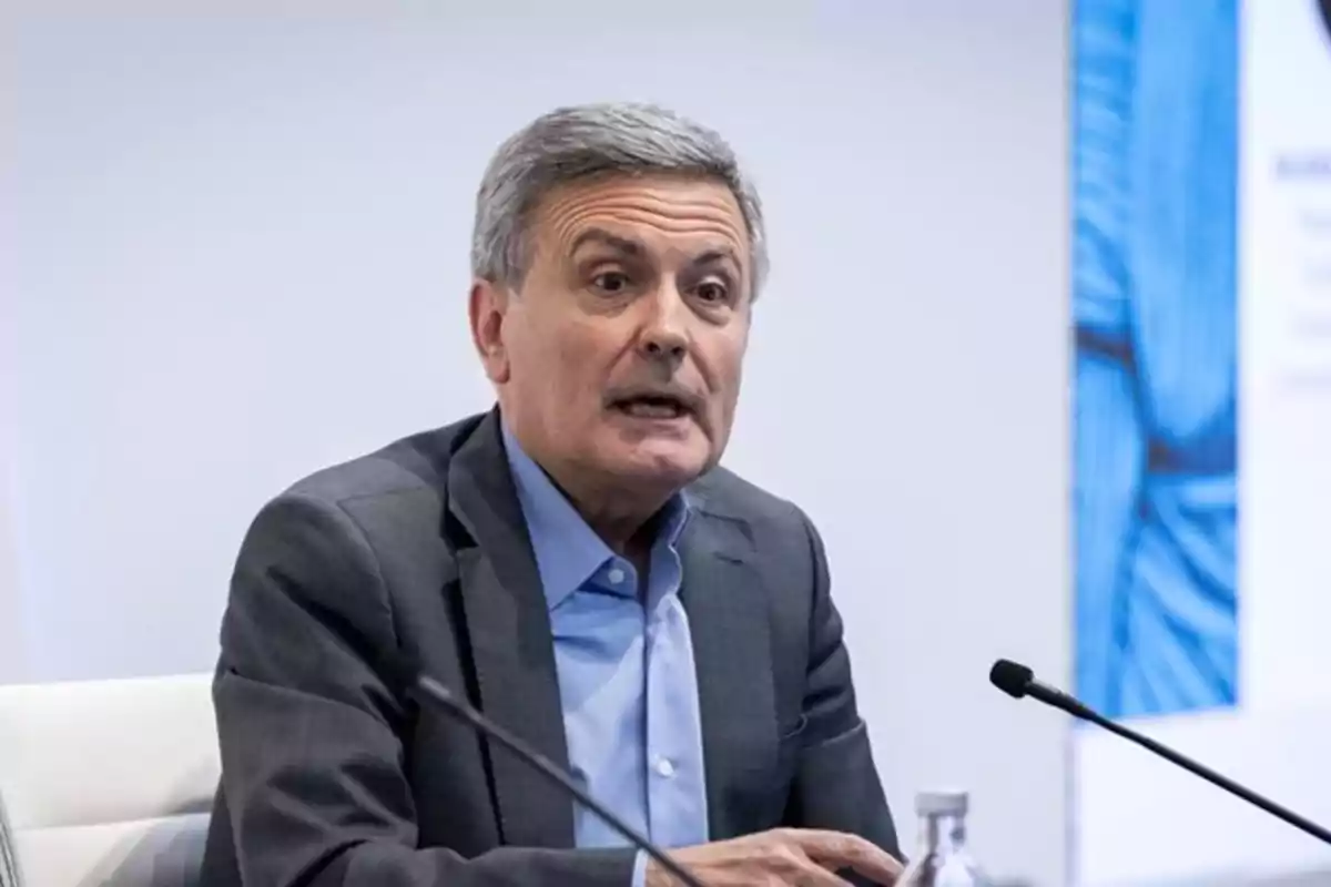 Hombre de cabello canoso con traje gris y camisa azul hablando frente a un micrófono en una sala de conferencias Hombre de cabello canoso con traje gris y camisa azul hablando frente a un micrófono en una sala de conferencias