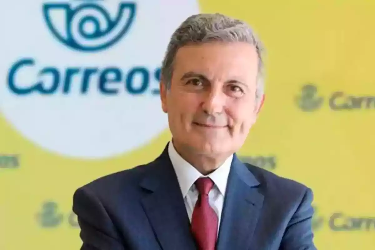 Hombre de cabello canoso con traje y corbata roja posando frente a un fondo amarillo con el logo y nombre de Correos