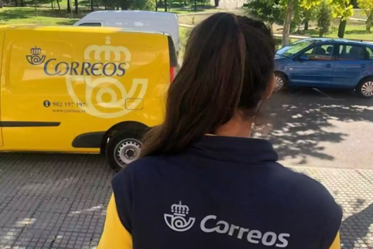 Una trabajadora de Correos de espaldas junto a una furgoneta amarilla de la empresa en la calle Una trabajadora de Correos de espaldas junto a una furgoneta amarilla de la empresa en la calle