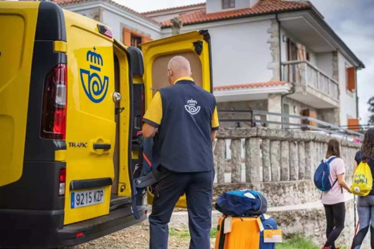Un trabajador de correos junto a una furgoneta amarilla abierta mientras entrega paquetes cerca de una casa y dos personas con mochilas pasan caminando Un trabajador de correos junto a una furgoneta amarilla abierta mientras entrega paquetes cerca de una casa y dos personas con mochilas pasan caminando