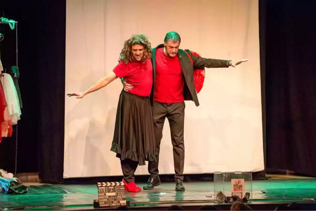 Dos actores en el escenario interpretan una escena cómica, ambos vestidos con camiseta roja, uno lleva falda y peluca mientras el otro usa saco y pantalón, con los brazos extendidos y una claqueta de cine en el suelo. Dos actores en el escenario interpretan una escena cómica, ambos vestidos con camiseta roja, uno lleva falda y peluca mientras el otro usa saco y pantalón, con los brazos extendidos y una claqueta de cine en el suelo.
