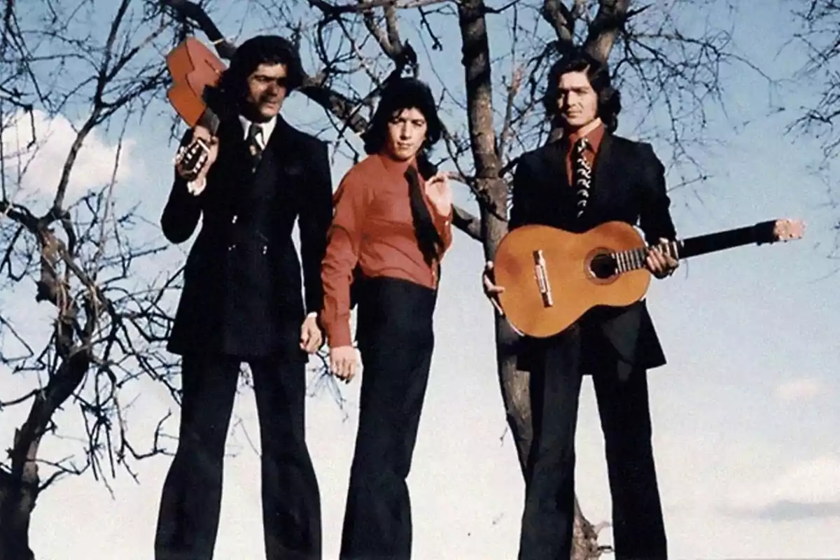 Tres hombres posan al aire libre junto a unos árboles sin hojas, dos de ellos sostienen guitarras y visten trajes formales de los años setenta Tres hombres posan al aire libre junto a unos árboles sin hojas, dos de ellos sostienen guitarras y visten trajes formales de los años setenta