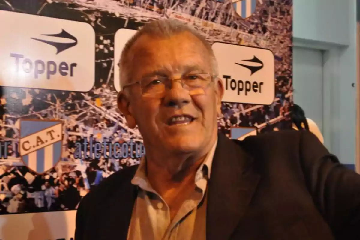 Hombre mayor con gafas y saco oscuro sonriendo frente a un fondo con logotipos de Topper y el escudo de un club de fútbol Hombre mayor con gafas y saco oscuro sonriendo frente a un fondo con logotipos de Topper y el escudo de un club de fútbol