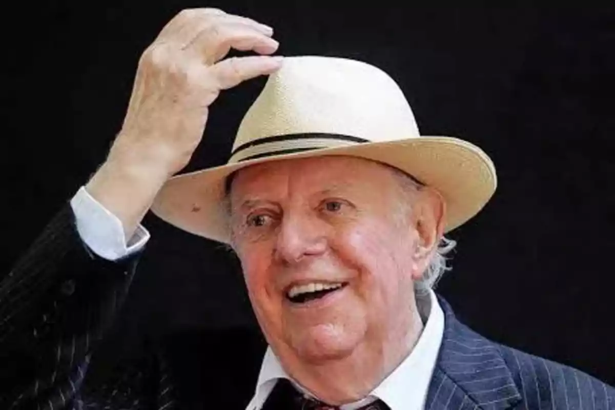 Hombre mayor sonriente con sombrero claro y traje oscuro tocándose la cabeza frente a un fondo negro