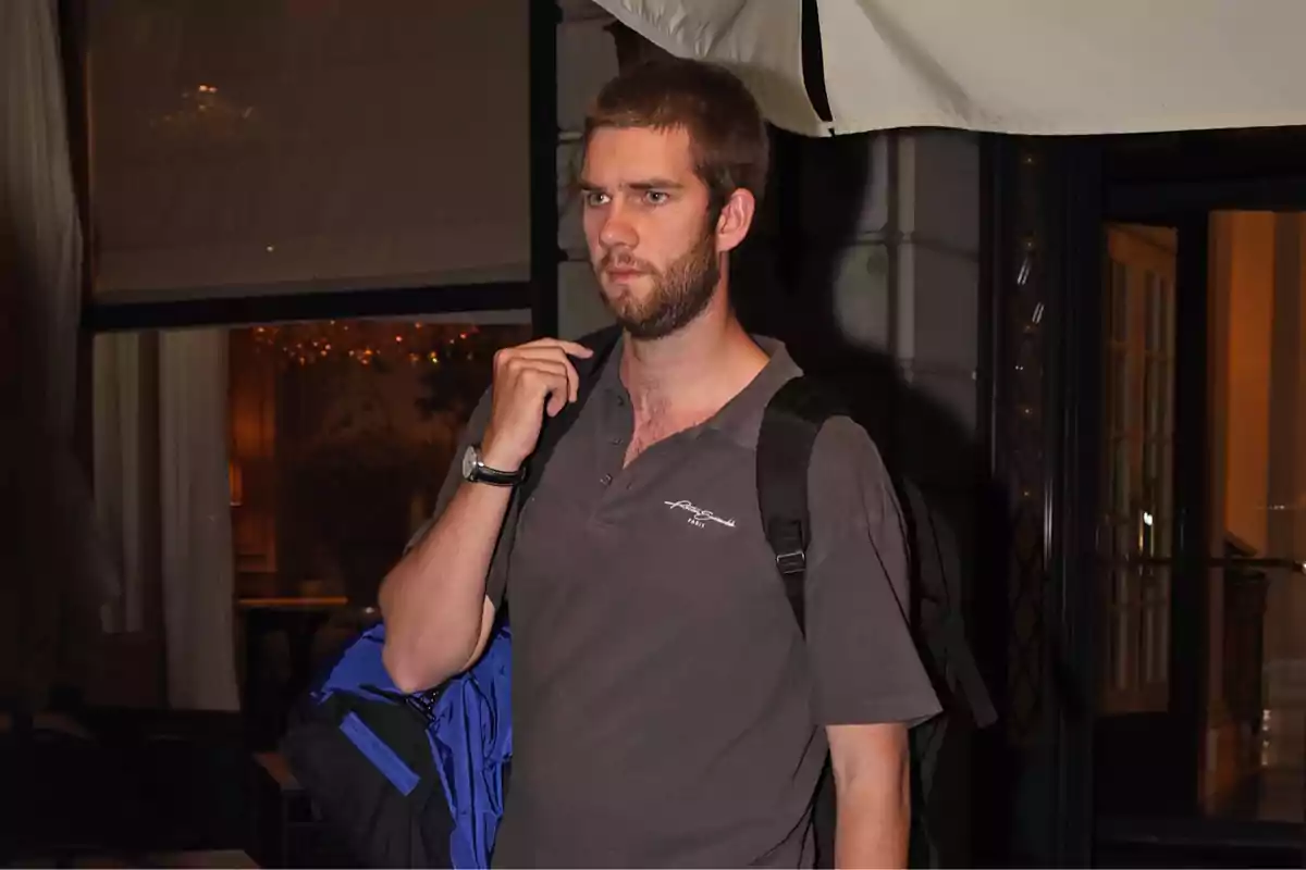 Young man with a beard and short hair carries a black backpack and wears a gray t-jersey while walking in an urban environment at night. Young man with a beard and short hair carries a black backpack and wears a gray t-jersey while walking in an urban environment at night.