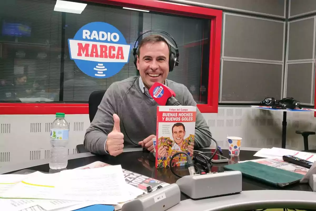 Hombre sonriente con auriculares y micrófono en un estudio de radio mostrando un libro titulado Buenas noches y buenos goles Hombre sonriente con auriculares y micrófono en un estudio de radio mostrando un libro titulado Buenas noches y buenos goles