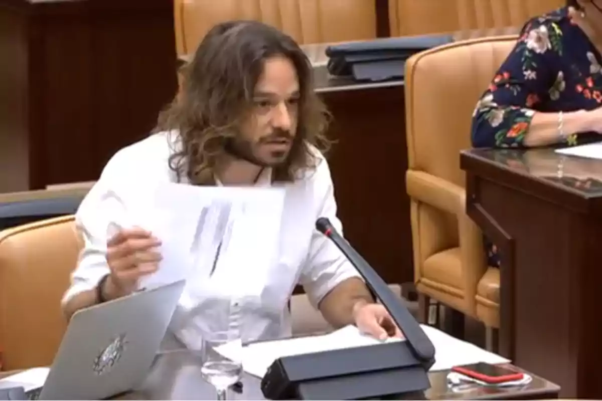 Hombre de cabello largo y barba hablando en una sala de reuniones mientras sostiene un documento frente a un micrófono Hombre de cabello largo y barba hablando en una sala de reuniones mientras sostiene un documento frente a un micrófono