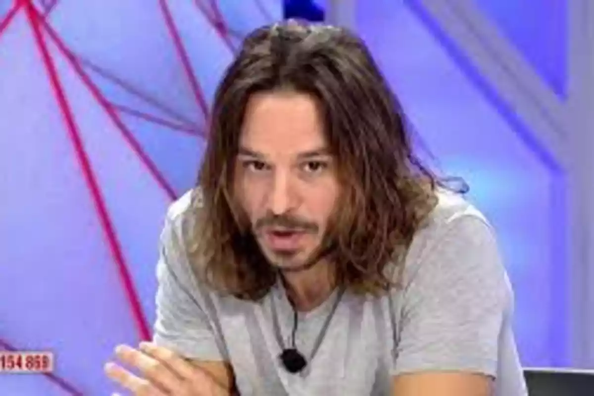 Hombre de cabello largo y barba usando camiseta gris y micrófono de solapa en un set de televisión con fondo azul Hombre de cabello largo y barba usando camiseta gris y micrófono de solapa en un set de televisión con fondo azul