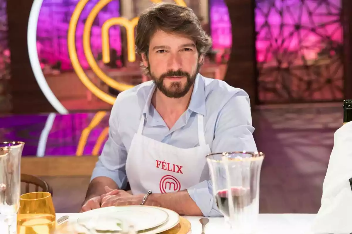 Hombre con barba y cabello castaño usando un delantal blanco con el nombre Félix sentado en una mesa de MasterChef con fondo colorido Hombre con barba y cabello castaño usando un delantal blanco con el nombre Félix sentado en una mesa de MasterChef con fondo colorido