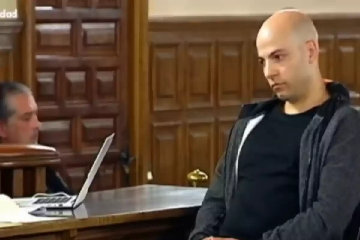 Hombre calvo con expresión seria sentado en una sala con paredes de madera y un hombre al fondo usando una laptop Hombre calvo con expresión seria sentado en una sala con paredes de madera y un hombre al fondo usando una laptop