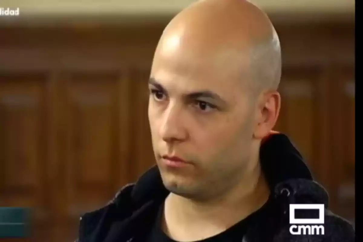 Hombre calvo con expresión seria vistiendo ropa oscura en un entorno interior con fondo de madera y logotipo de CMM en la esquina inferior derecha Hombre calvo con expresión seria vistiendo ropa oscura en un entorno interior con fondo de madera y logotipo de CMM en la esquina inferior derecha