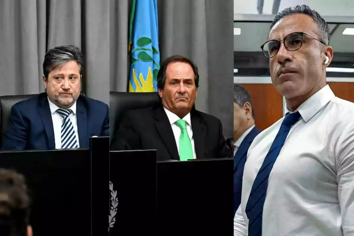 Dos hombres con traje y corbata sentados en un estrado junto a una bandera y un hombre de pie con camisa blanca y corbata azul mirando hacia el frente Dos hombres con traje y corbata sentados en un estrado junto a una bandera y un hombre de pie con camisa blanca y corbata azul mirando hacia el frente