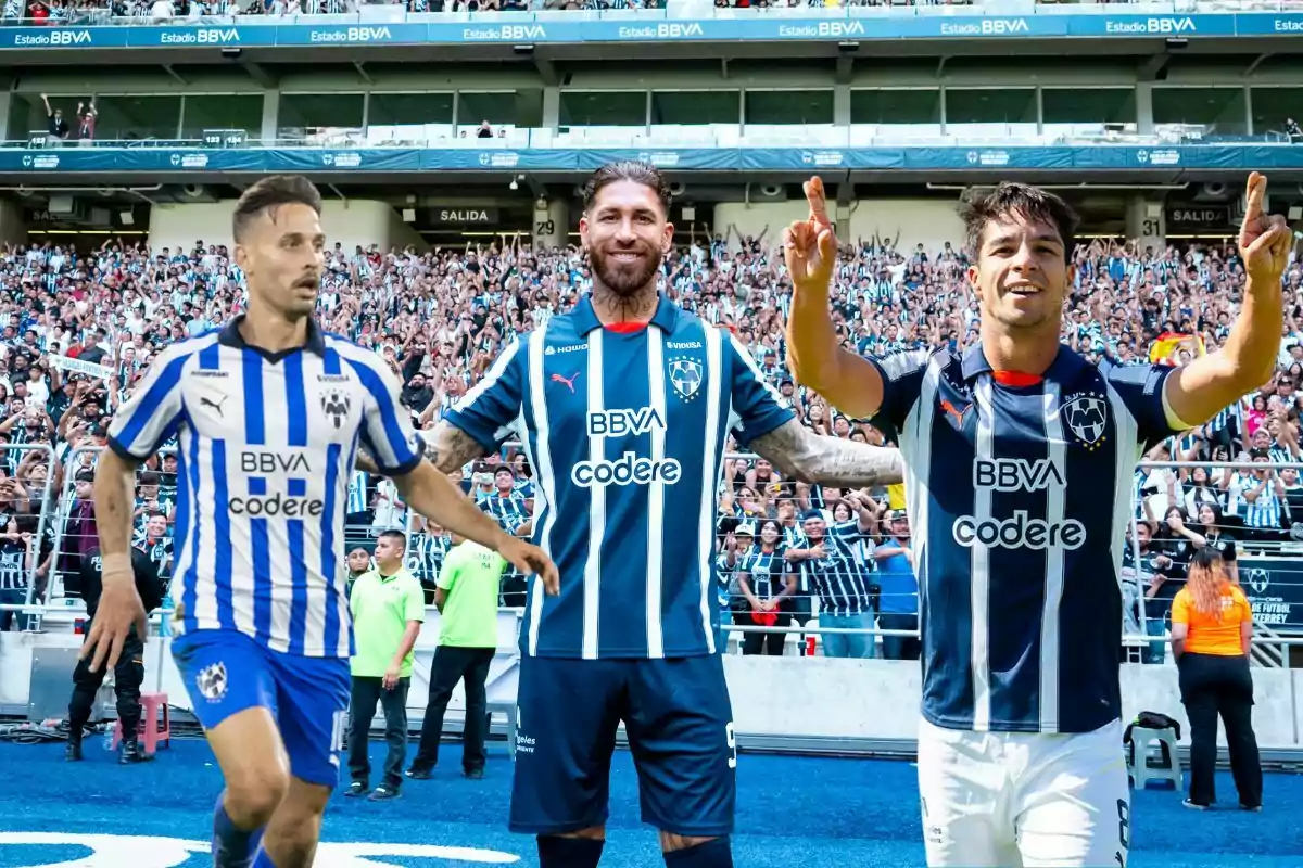 Tres futbolistas con uniforme de Rayados de Monterrey celebran en la cancha frente a una multitud de aficionados en el estadio BBVA