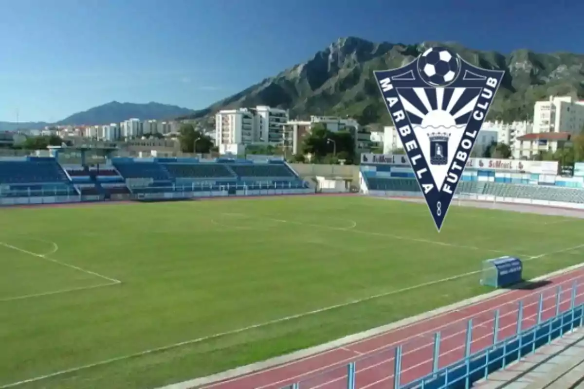 Estadio de fútbol con gradas azules, pista de atletismo y el escudo del Marbella Fútbol Club sobrepuesto, con montañas y edificios al fondo