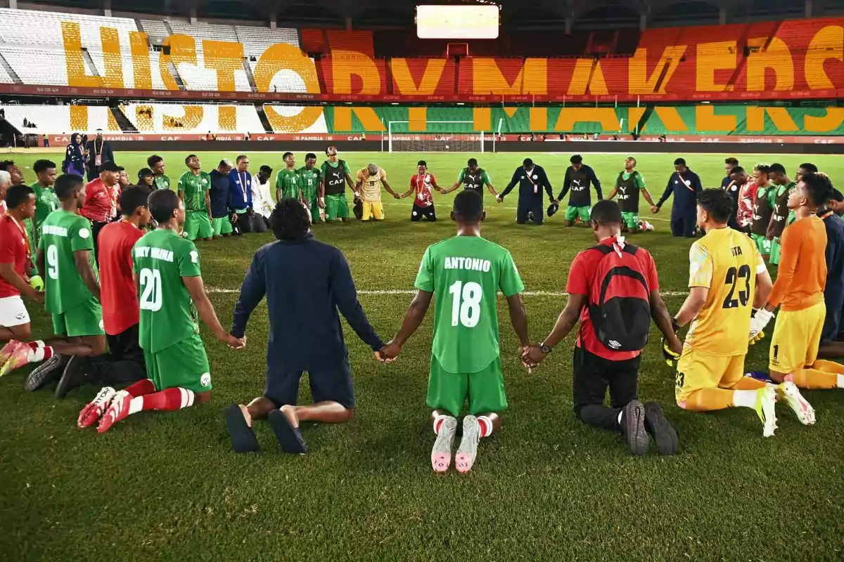 Jugadores de fútbol arrodillados en círculo tomados de las manos en el campo, con la palabra HISTORY MAKERS en las gradas del estadio
