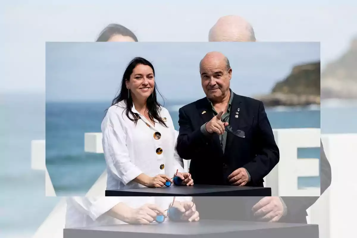 Dos personas posan sonrientes al aire libre frente al mar una de ellas viste una camisa blanca y sostiene unos lentes mientras la otra persona señala hacia la cámara