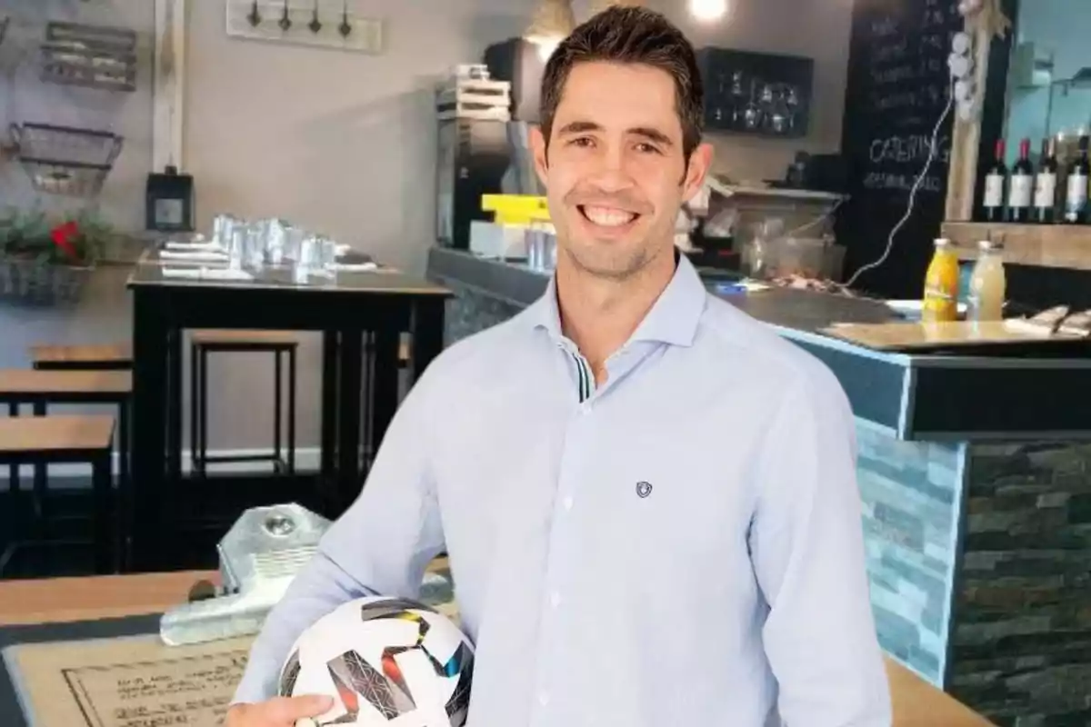 Hombre sonriente con camisa azul claro sosteniendo un balón de fútbol en el interior de un restaurante moderno