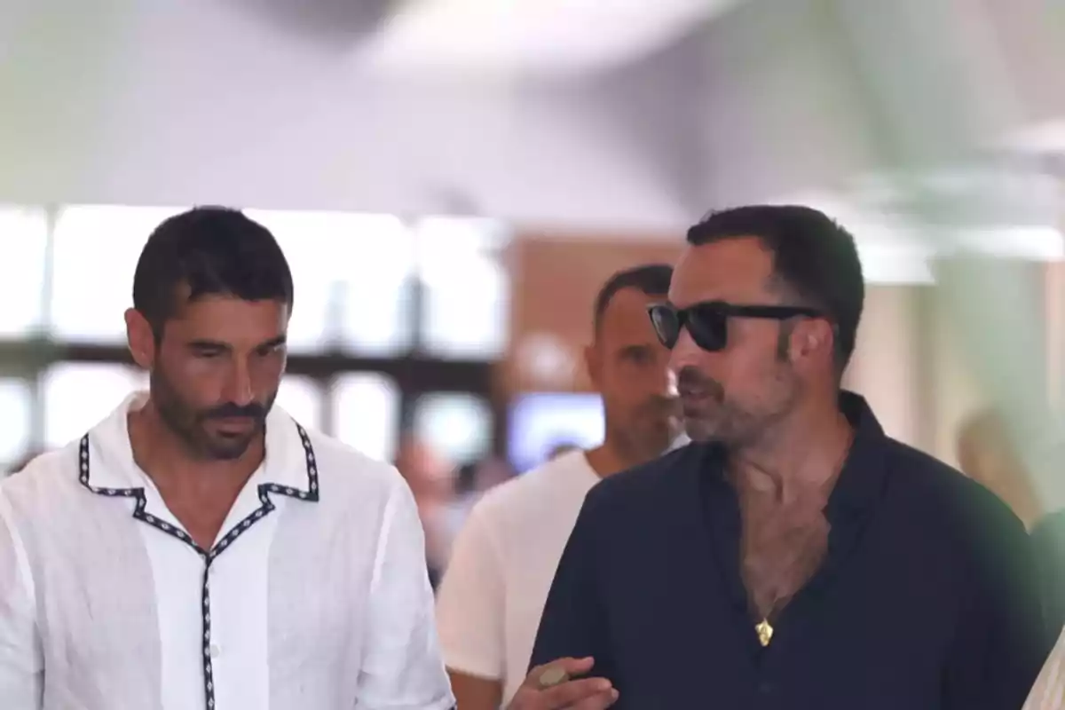 Dos hombres caminan juntos en un lugar cerrado mientras conversan, uno lleva camisa blanca y el otro camisa oscura y gafas de sol Dos hombres caminan juntos en un lugar cerrado mientras conversan, uno lleva camisa blanca y el otro camisa oscura y gafas de sol
