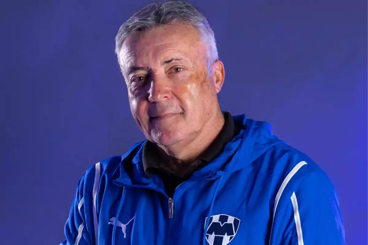 Hombre de cabello canoso vistiendo una chaqueta azul con el escudo del club de fútbol Monterrey y el logo de Puma sobre un fondo azul Hombre de cabello canoso vistiendo una chaqueta azul con el escudo del club de fútbol Monterrey y el logo de Puma sobre un fondo azul