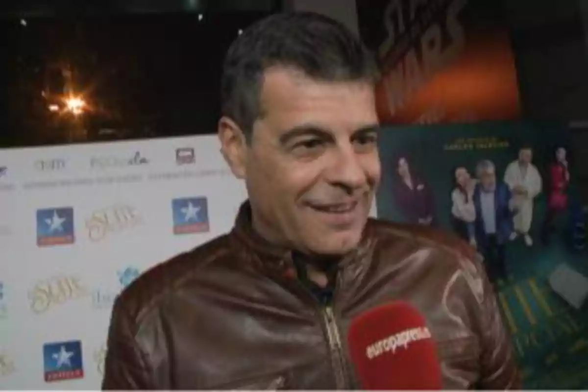 Hombre con chaqueta de cuero marrón sonriendo mientras sostiene un micrófono rojo en un evento con carteles de fondo Hombre con chaqueta de cuero marrón sonriendo mientras sostiene un micrófono rojo en un evento con carteles de fondo
