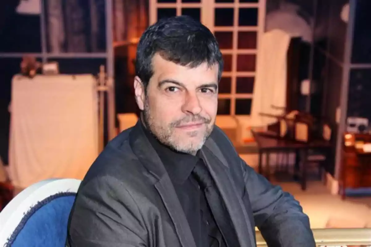 Hombre de cabello corto y barba ligera con traje oscuro sentado en una silla azul en un ambiente interior elegante