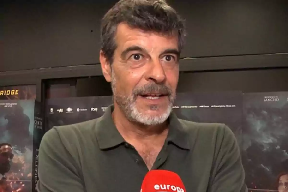 Hombre de cabello canoso y barba con camisa polo verde oscuro sosteniendo un micrófono rojo en un evento de prensa con carteles de fondo Hombre de cabello canoso y barba con camisa polo verde oscuro sosteniendo un micrófono rojo en un evento de prensa con carteles de fondo