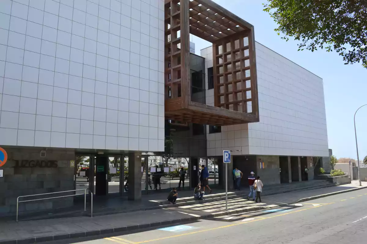 Edificio moderno de juzgados con fachada de paneles blancos y una estructura de madera en forma de cubo suspendida sobre la entrada principal donde varias personas caminan y esperan en la acera Edificio moderno de juzgados con fachada de paneles blancos y una estructura de madera en forma de cubo suspendida sobre la entrada principal donde varias personas caminan y esperan en la acera