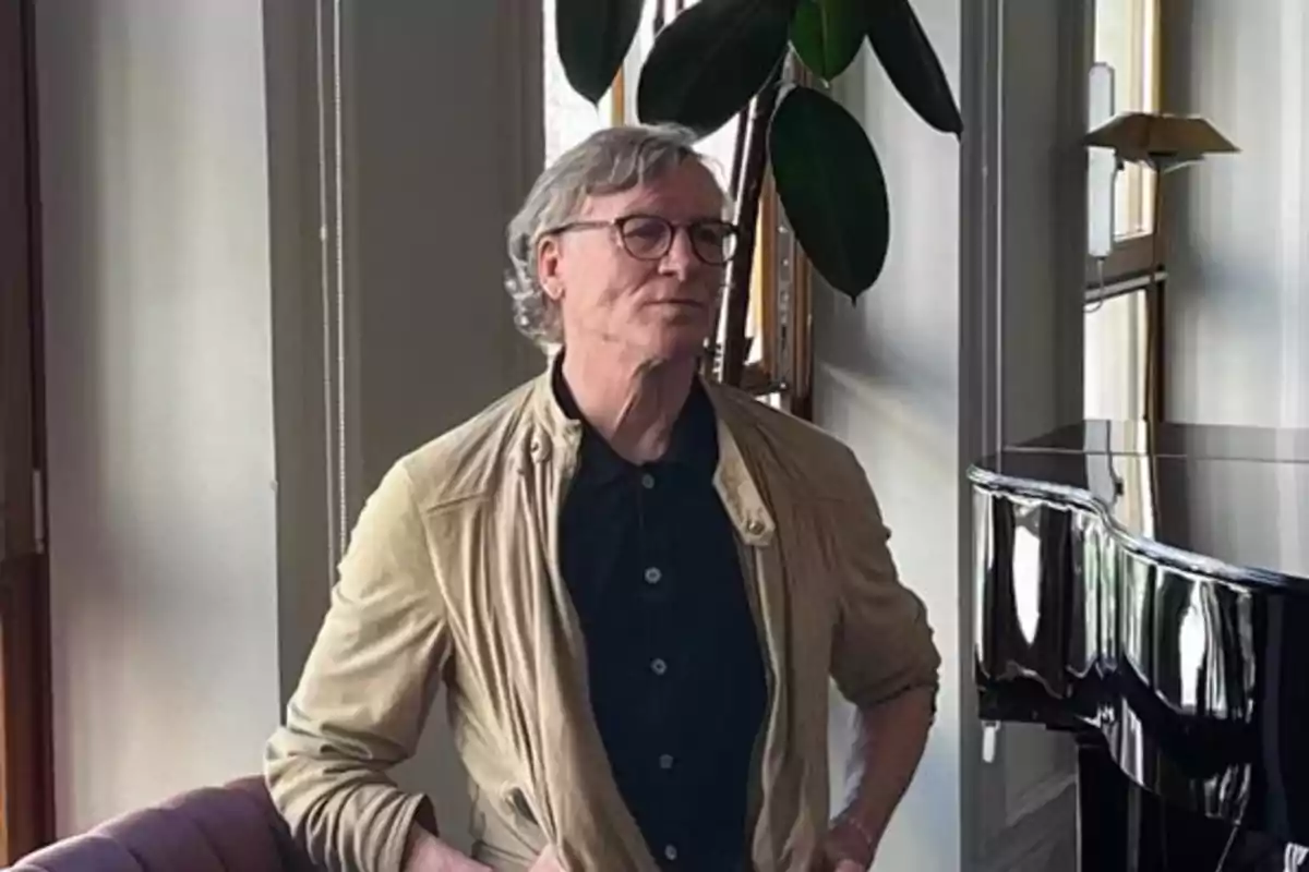 Hombre de cabello canoso y gafas de pie junto a un piano en una habitación iluminada Hombre de cabello canoso y gafas de pie junto a un piano en una habitación iluminada